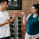 Side view of Asian personal instructor with apple and doughnut offering healthy food to plus size woman while standing in gym